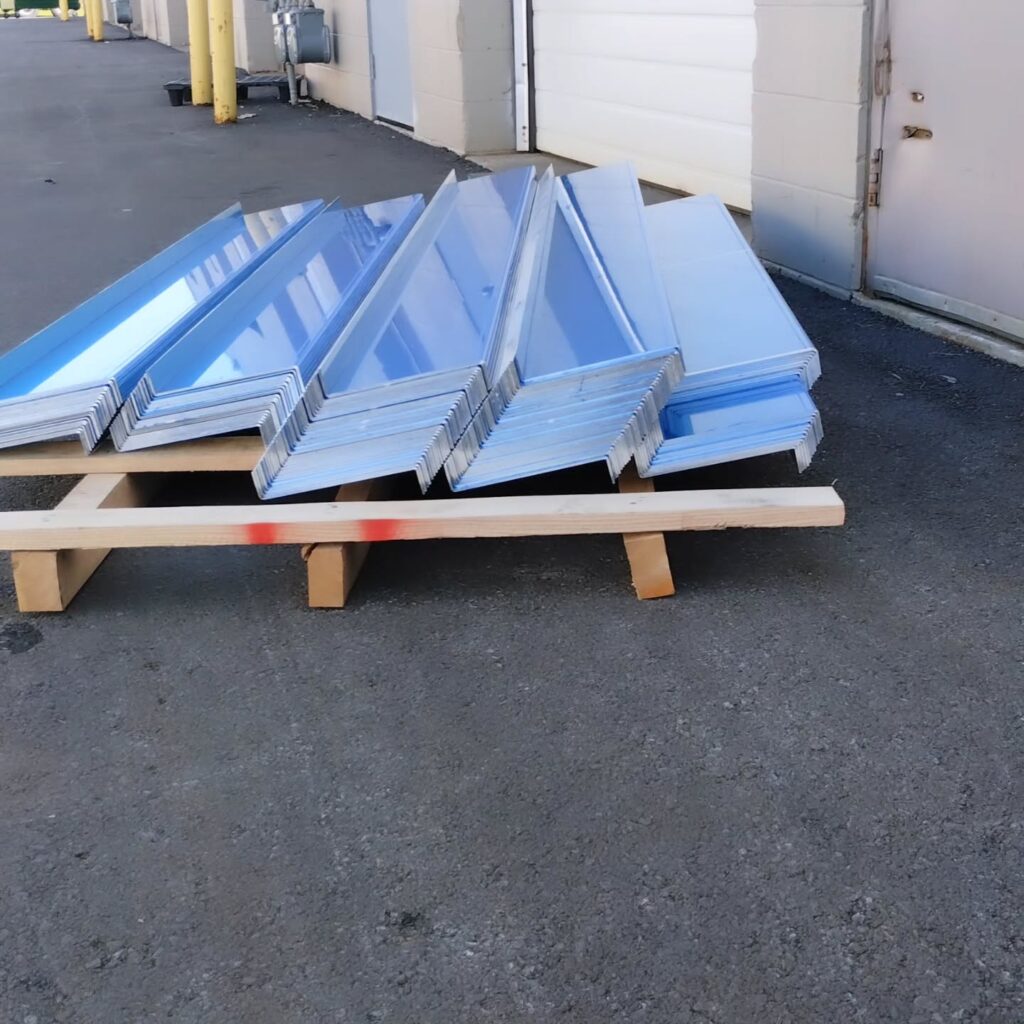 Metal sheets stacked on a wooden pallet outside a building, reflecting sunlight. These sheets are likely materials for construction or manufacturing.
