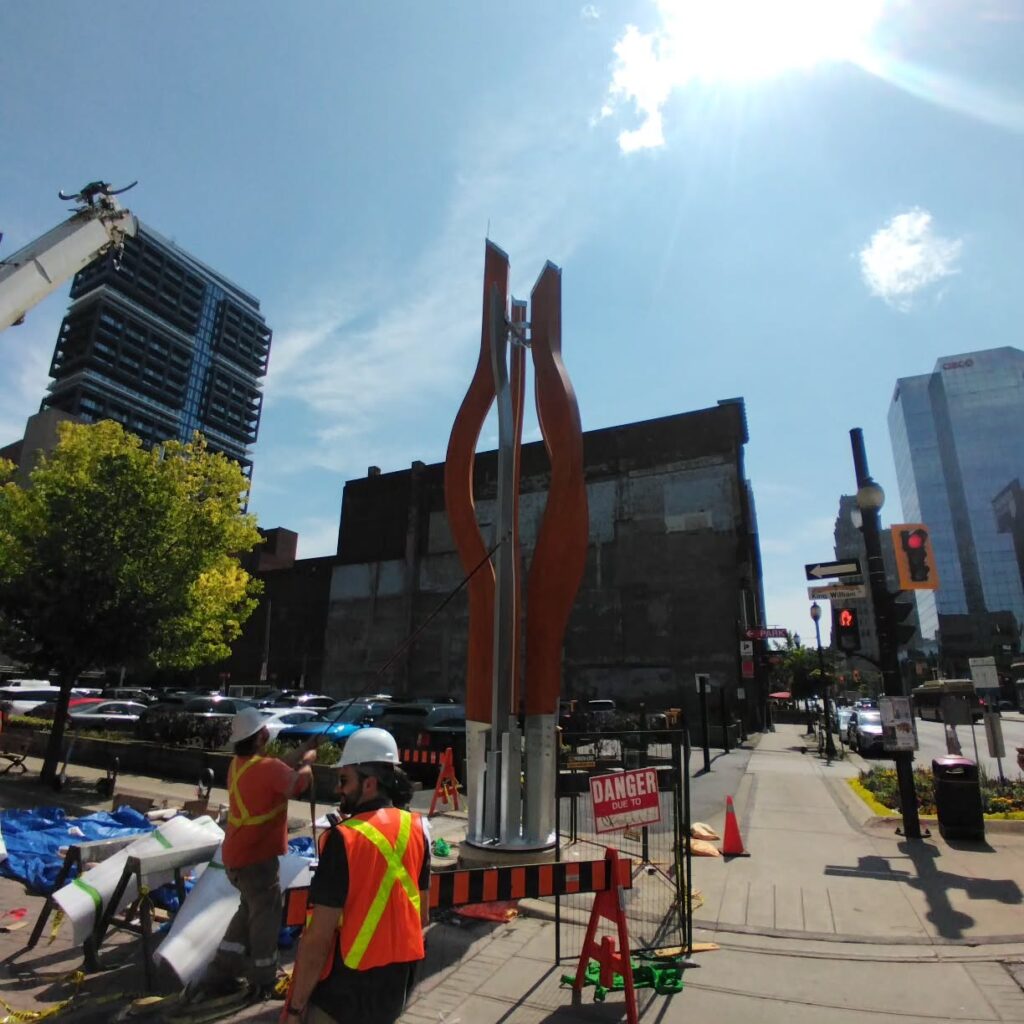 Construction workers install a large, abstract metal sculpture in an urban setting, with buildings and traffic in the background.