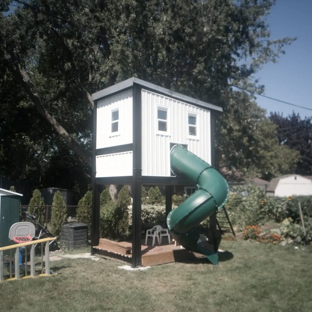 Elevated playhouse with a green slide in a backyard. Features include large windows and a wooden deck, offering a fun play space for children.
