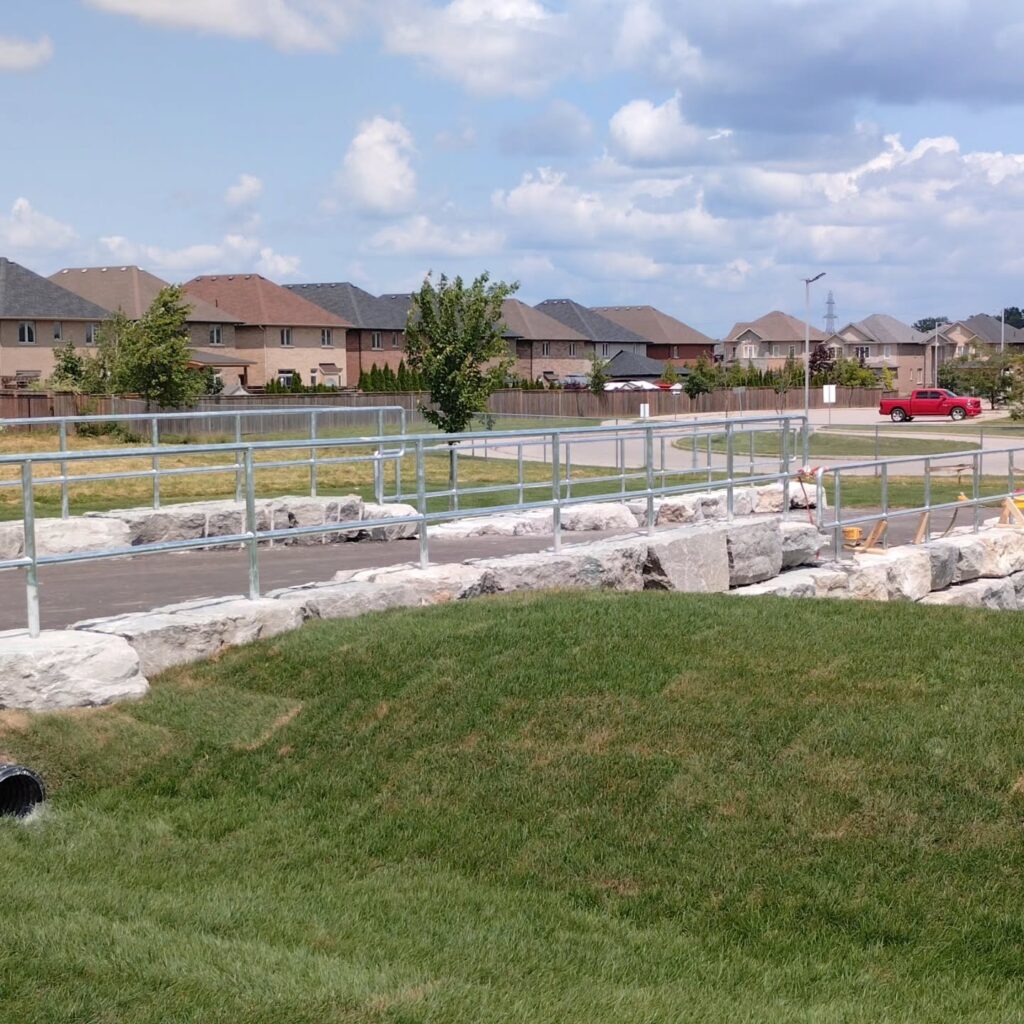 Newly constructed pathway with metal railing over rocky terrain, surrounded by green grass and residential homes under a partly cloudy sky.