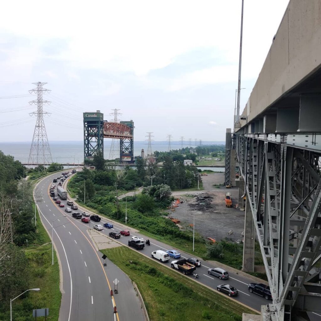 Traffic flows along a winding road beneath a large, elevated bridge featuring a "Canada" sign, overlooking a body of water and power lines.