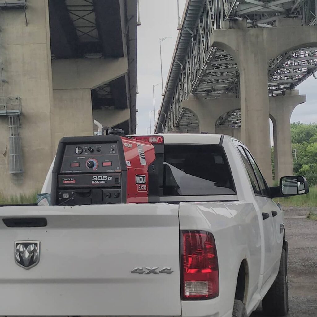 A white pickup truck carries a Lincoln Electric Ranger 305G welder in its bed, parked under a concrete overpass surrounded by greenery.