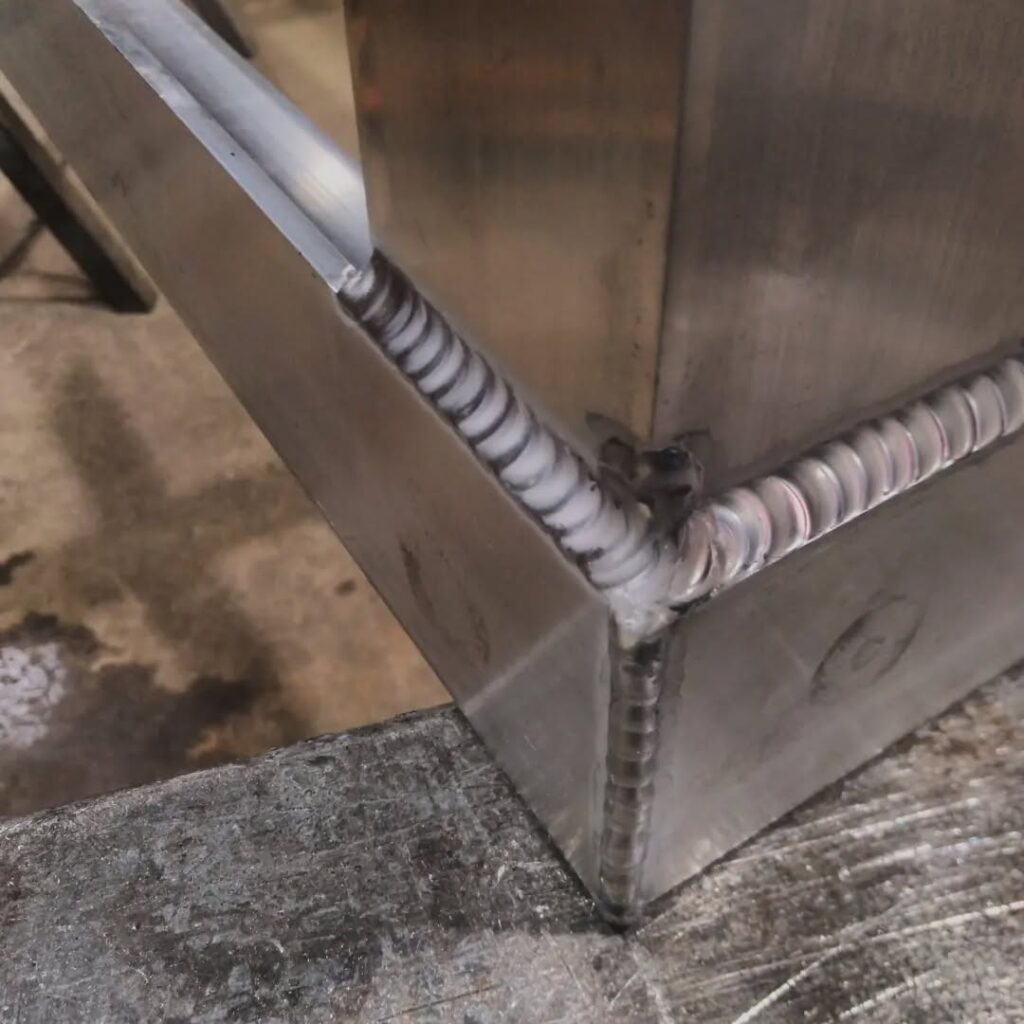 Close-up of a clean aluminum weld joint, showcasing precise bead patterns and the corner of a fabricated metal piece, highlighting welding quality.