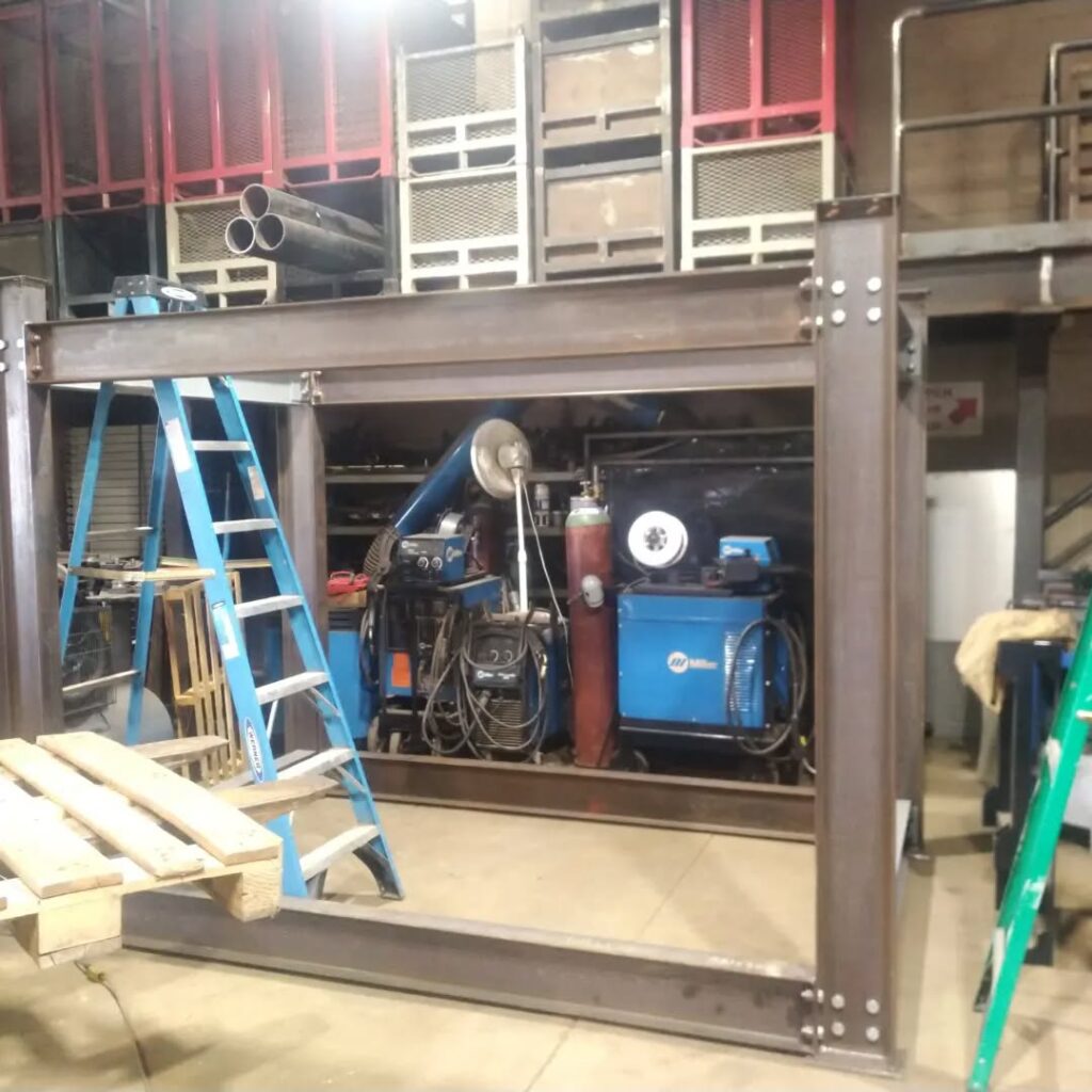 A steel frame structure in a workshop, with a blue ladder and equipment in the background, illustrating a workspace for fabrication or welding.