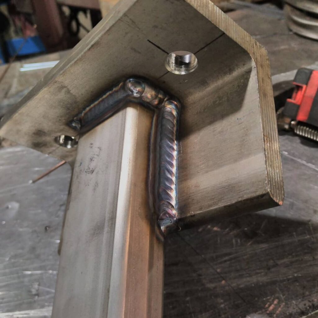 Close-up of a welded metal joint, showcasing intricate weld patterns on a steel bracket. The image highlights the craftsmanship in metal fabrication.