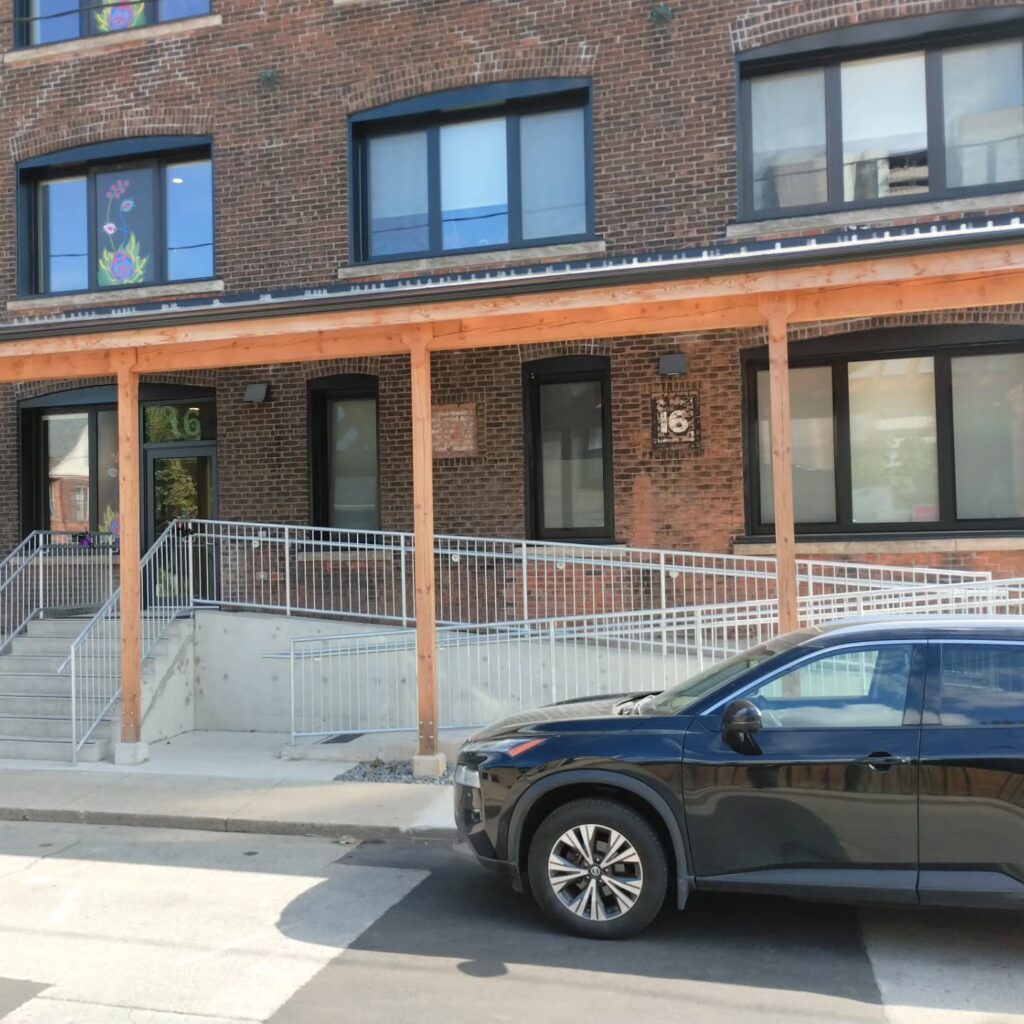 Modern brick building with a wooden overhang, featuring an accessible ramp. A black SUV is parked nearby, indicating a residential area.