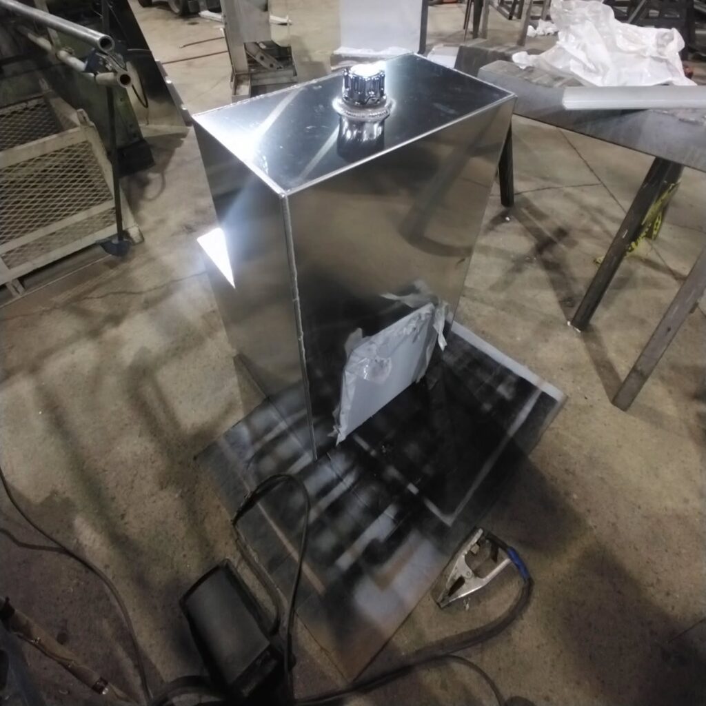 Shiny stainless steel box with a vent on a workshop floor, showcasing a fabrication project in progress. Tools and materials are visible in the background.