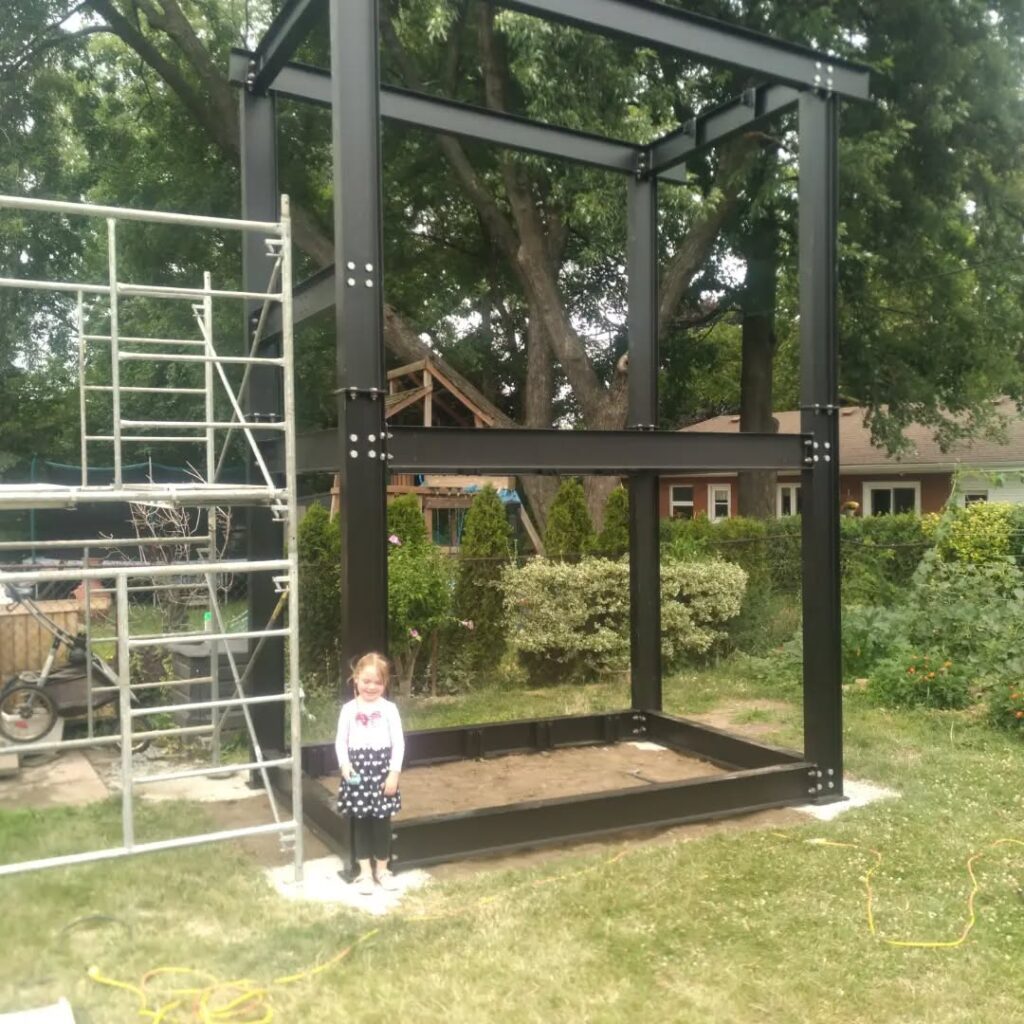 Child standing next to an unfinished metal play structure in a backyard, with scaffolding visible nearby. The scene captures ongoing construction.