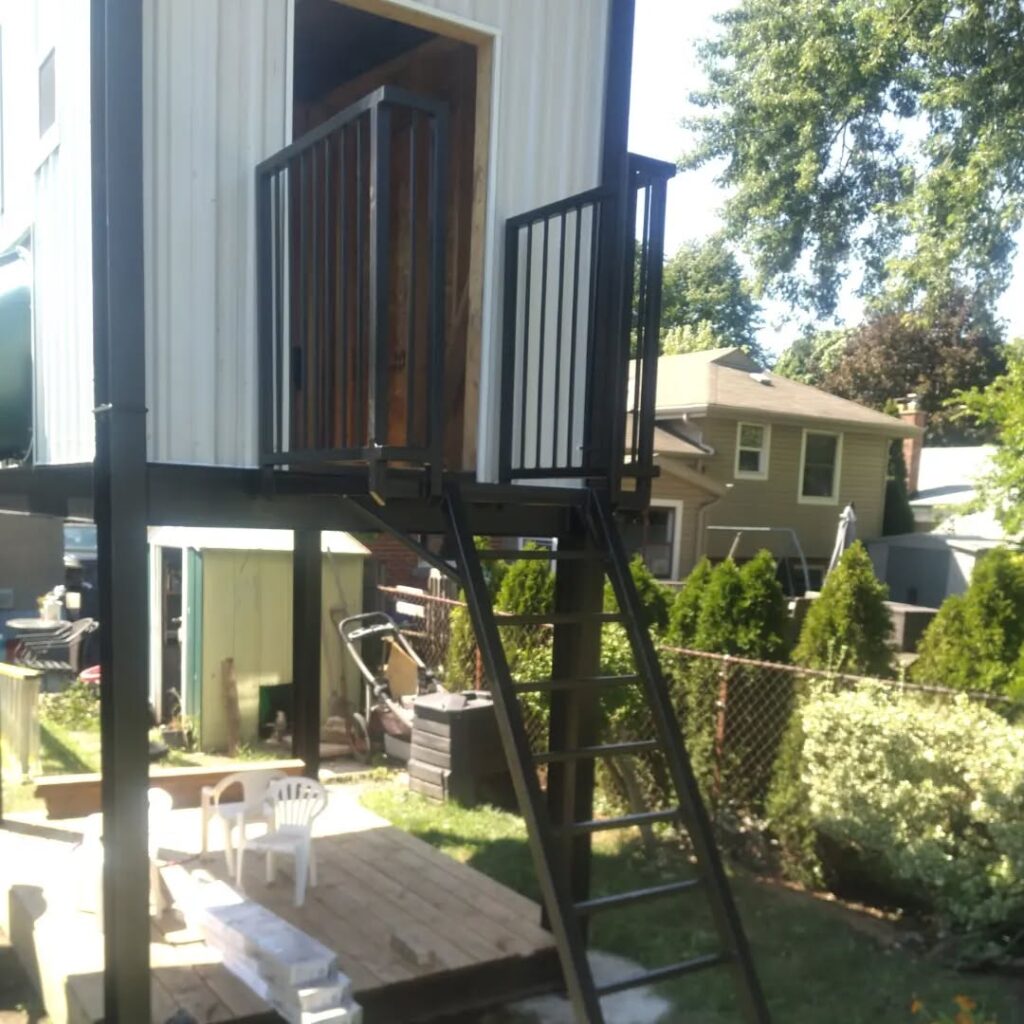 Elevated playhouse with a metal staircase and balcony, situated in a backyard surrounded by greenery and outdoor furniture.