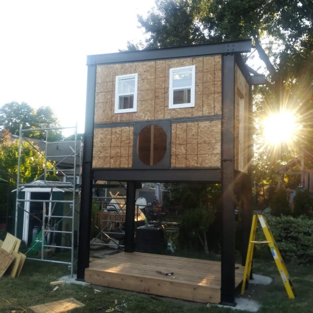 A partially constructed playhouse with two windows and a circular opening. Surrounding scaffolding and tools indicate ongoing construction in a backyard.