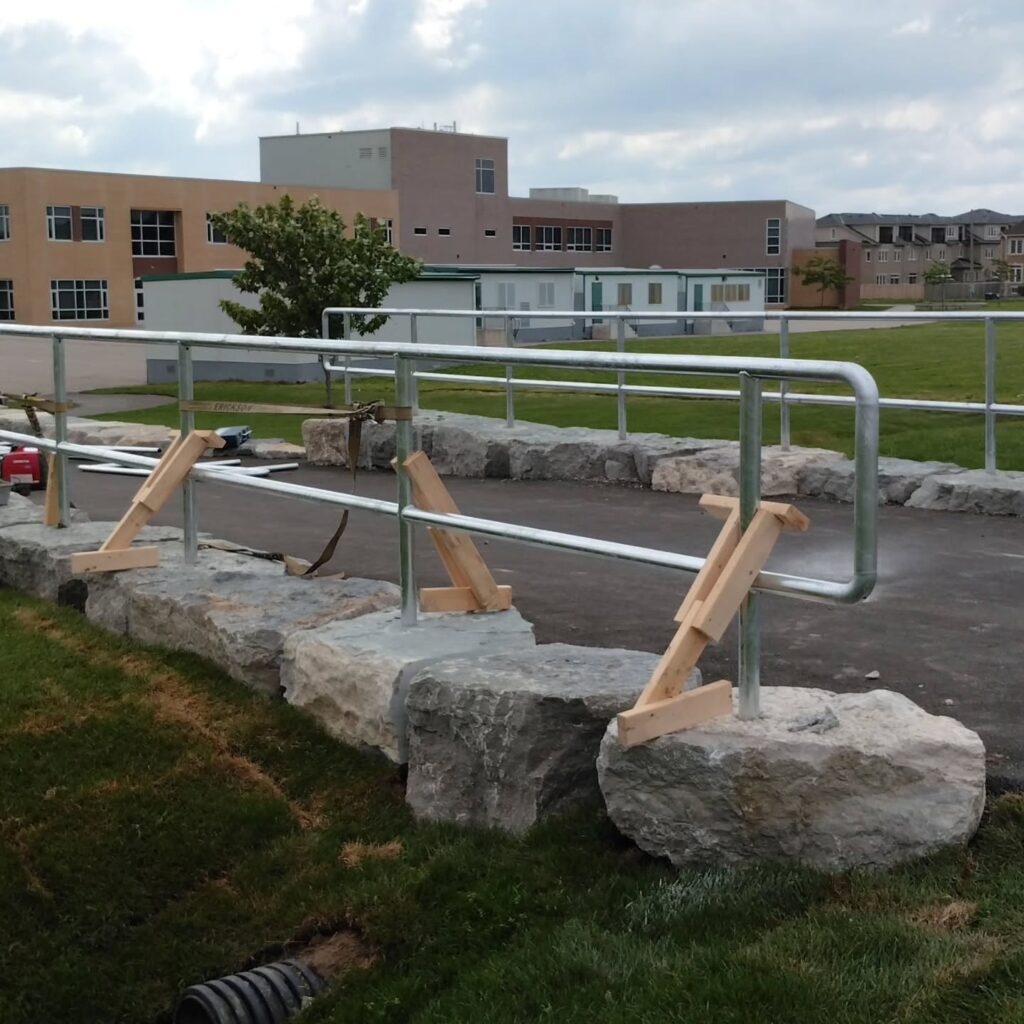 A wooden support structure stabilizes a metal railing on a pathway near a school building, illustrating ongoing outdoor construction or maintenance.
