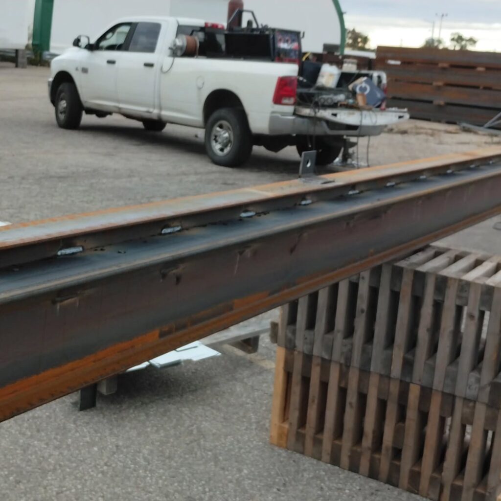 Steel beam positioned for loading with a white truck in the background. The image shows a construction or industrial setting.
