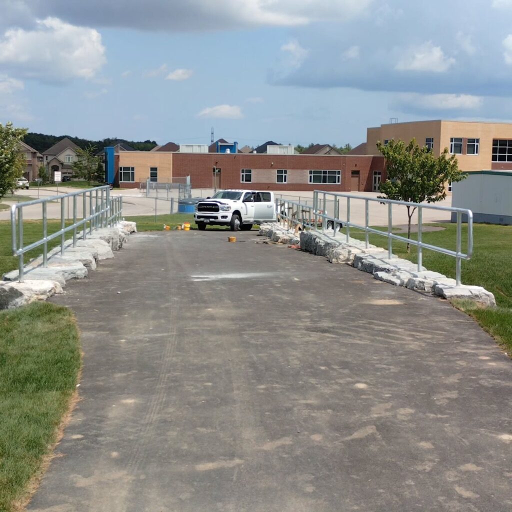 Newly constructed pathway leading to a school, featuring a concrete surface and rock barriers, with a white truck parked nearby.