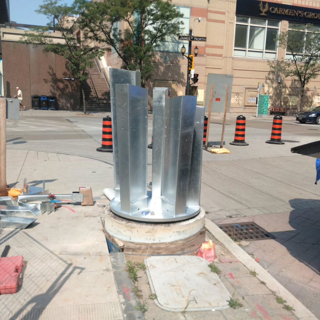 Metal construction elements are installed in a concrete base on a street corner, surrounded by construction barriers and signage.
