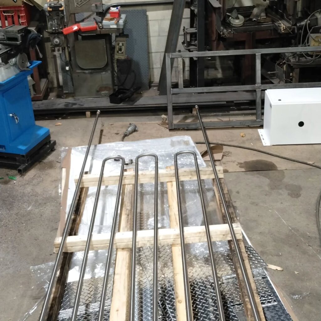 Metal rods arranged on a wooden pallet in a workshop, surrounded by machinery and tools, highlighting an industrial fabrication setting.