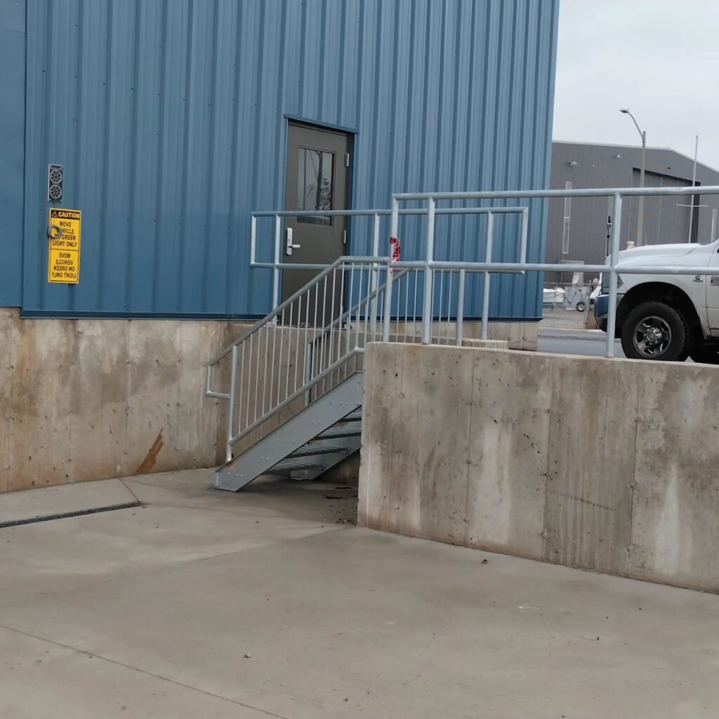 Metal staircase leading to a blue metal door, with a caution sign visible nearby. Concrete barrier and a white truck are in the foreground.