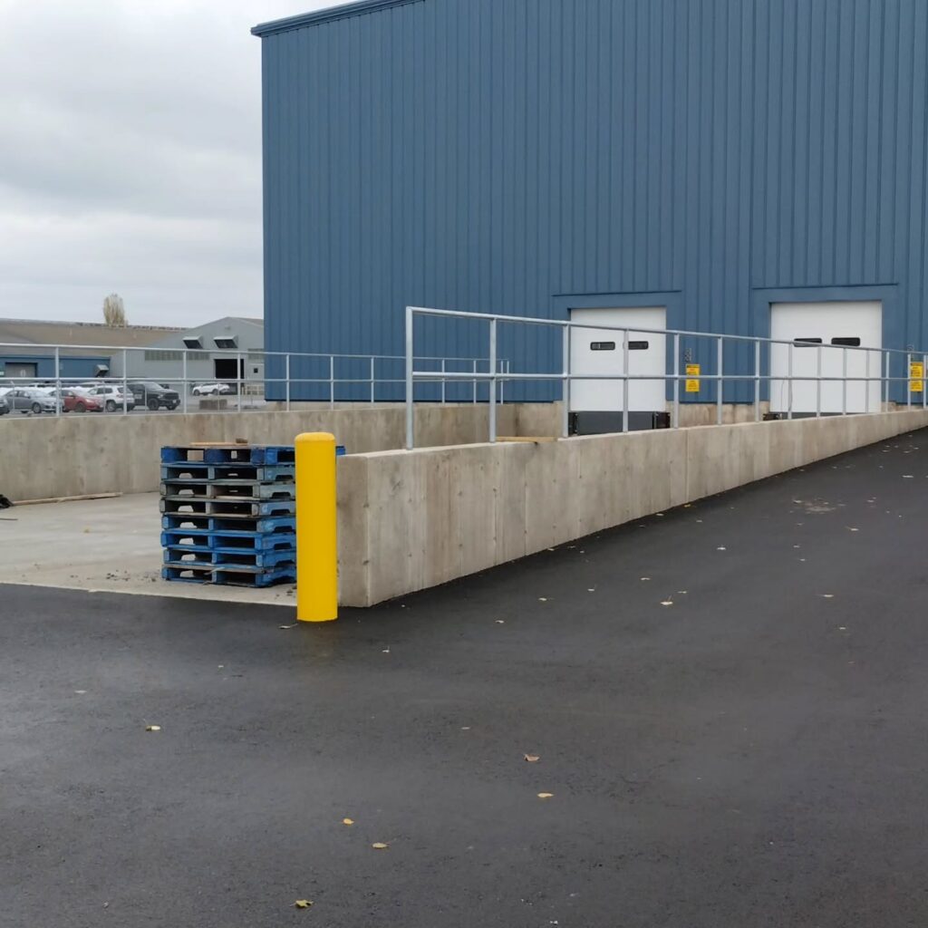 Loading dock area with concrete barrier, yellow post, and stacked pallets in front of a blue industrial building. Ideal for freight operations.