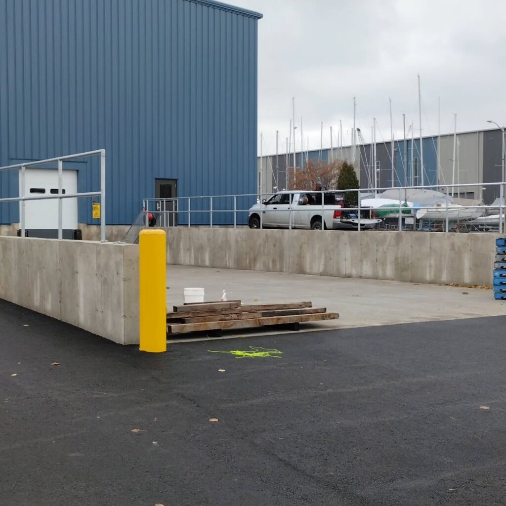 Concrete loading area beside a blue metal building, with a white pickup truck parked nearby and sailboats visible in the background.
