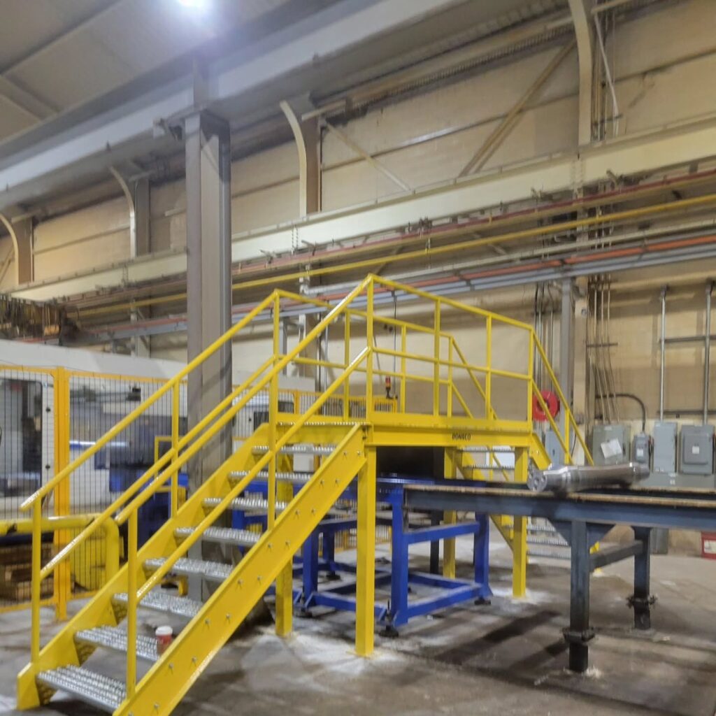 Yellow metal stairs lead to a platform in a spacious industrial setting, with machinery and electrical panels in the background.