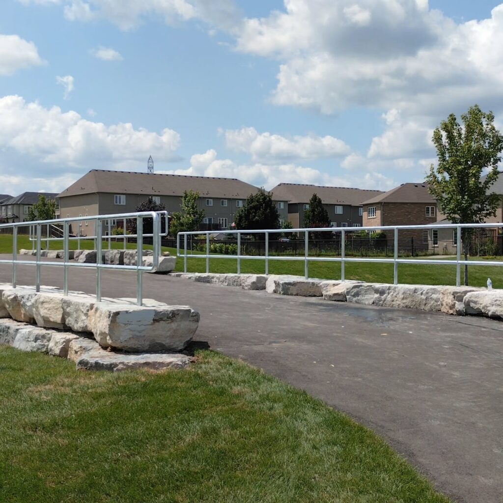 A wide paved path with a railing curves over large rocks, leading to houses in a suburban area under a bright blue sky with clouds.