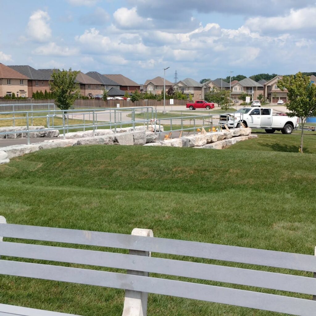 Landscape view of a park area featuring a grassy hill, a stone arrangement, and a white pickup truck nearby. Residential homes and cloudy sky in the background.