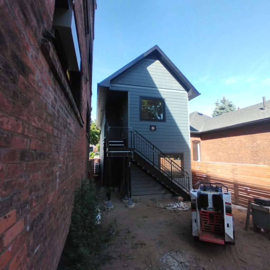 Newly constructed gray house with a gabled roof, featuring a metal staircase on the side. Construction materials are nearby, indicating ongoing work.
