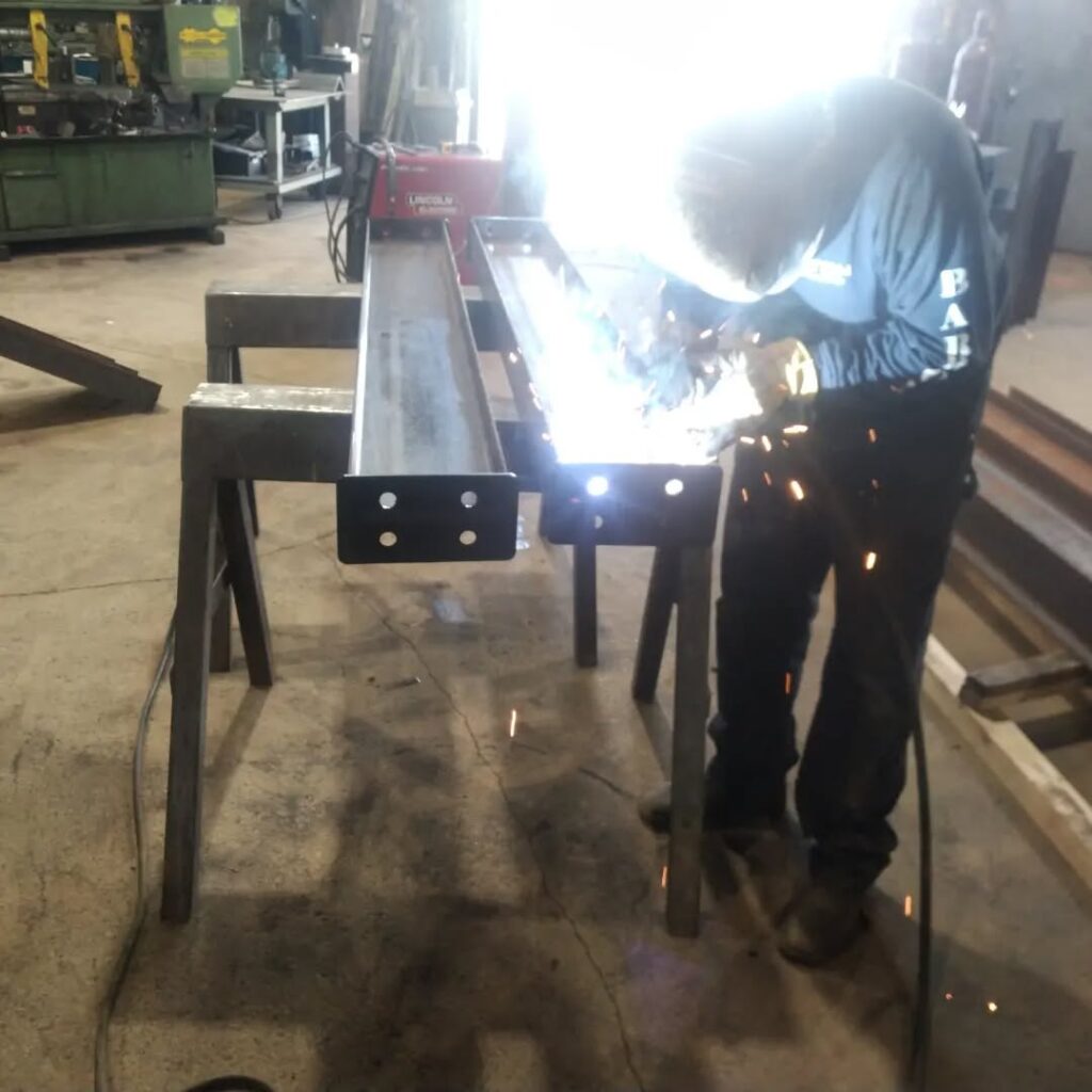 A welder fuses two metal beams in a workshop, producing sparks and bright light, showcasing metal fabrication techniques.