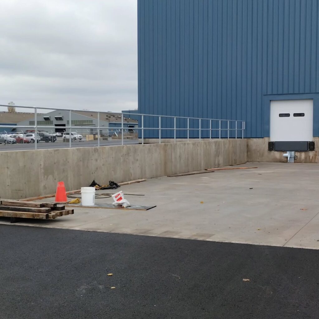 Loading dock area with a blue metal building, concrete ramp, orange cone, and scattered construction materials. It highlights a workspace readiness.