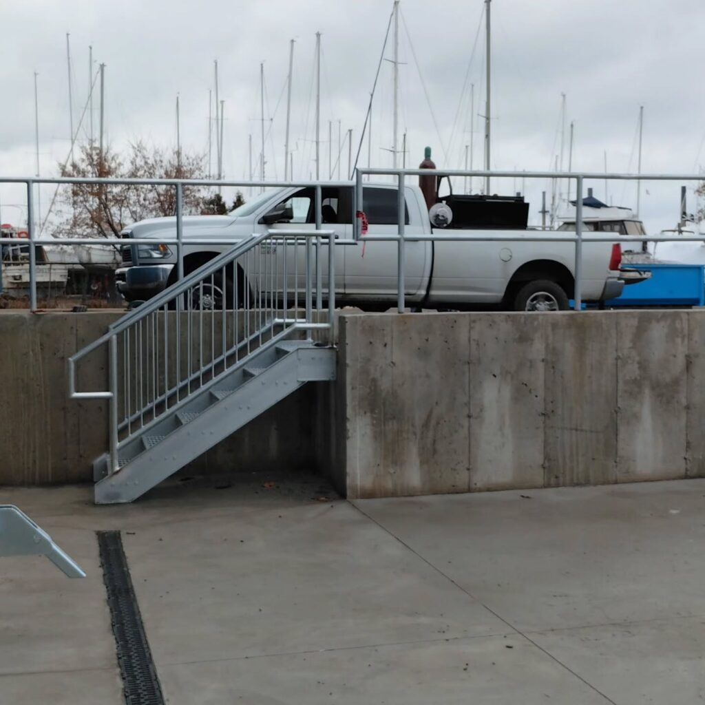 White pickup truck parked next to a steel staircase leading to a marina. Sailboats and a cloudy sky in the background reflect a coastal setting.