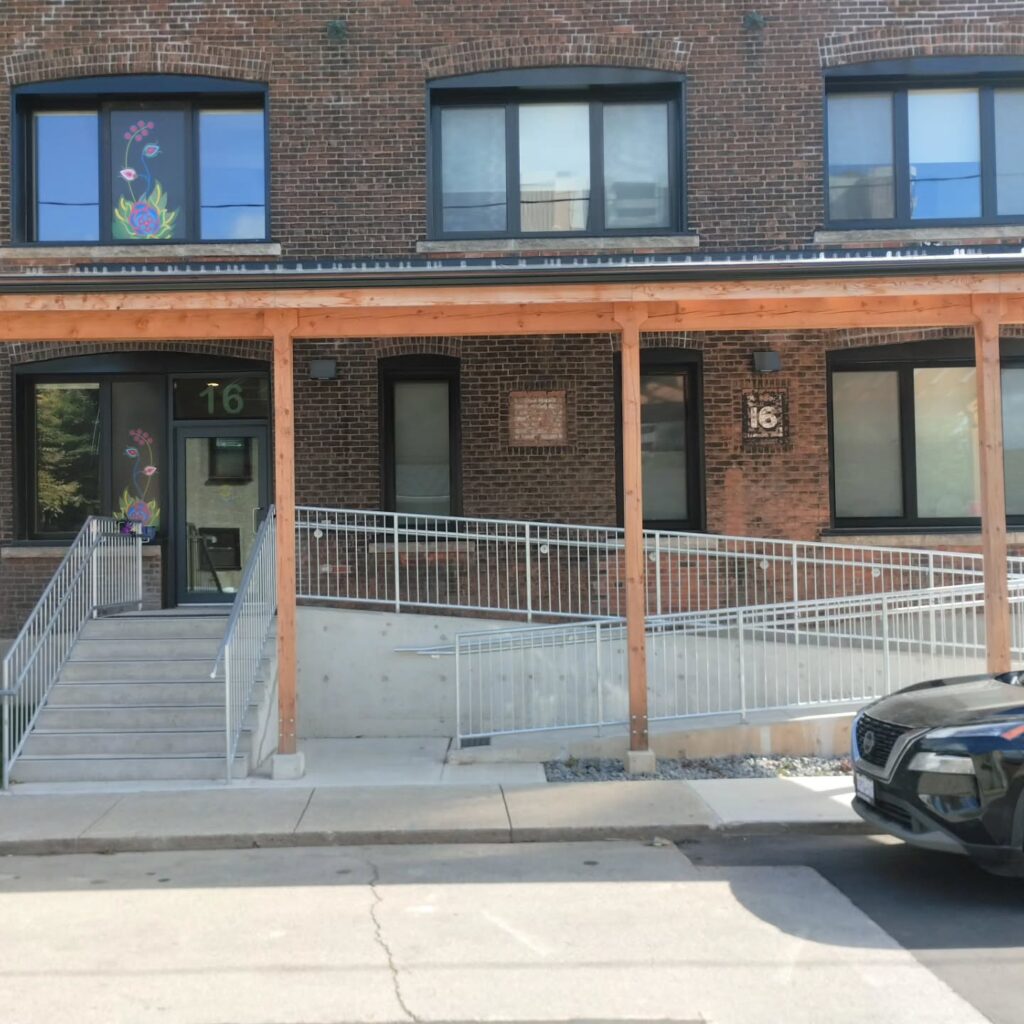 Brick building entrance with a prominent رقم 16. Features a decorated door and accessible ramp, surrounded by modern architecture.