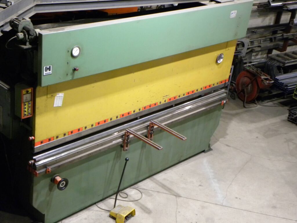 Hydraulic bending machine in a workshop, featuring a green and yellow exterior, control panel, and metal sheets ready for processing.
