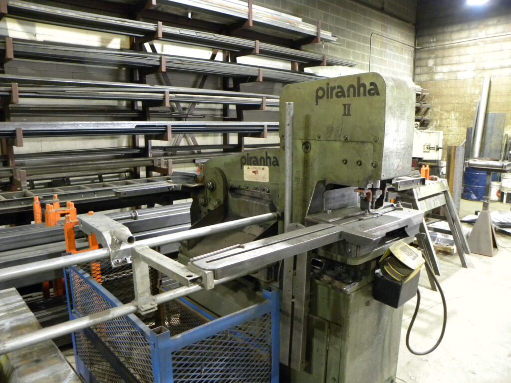 Piranha II metal shear machine in a workshop, surrounded by various steel beams and tools, illustrating industrial manufacturing processes.